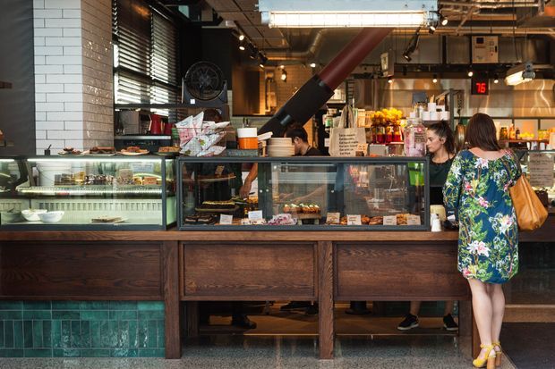 Interior view of a casual café or food-service counter with a long dark timber display cabinet, glass pastry and food cases, tiled feature wall, exposed industrial-style ceiling services, pendant lighting, and staff serving customers. The space combines warm timber joinery with teal-green wall tiles and utilitarian commercial detailing.