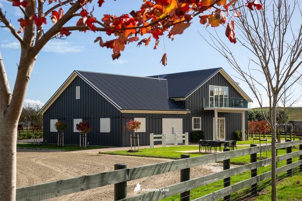 Exterior view of a contemporary dark-clad house with multiple pitched gables, standing behind a timber post-and-rail fence. The building has vertical dark weatherboard-style cladding, a dark metal roof, white-framed windows, and landscaped grounds with a gravel driveway and young trees. Autumn foliage frames the upper foreground.