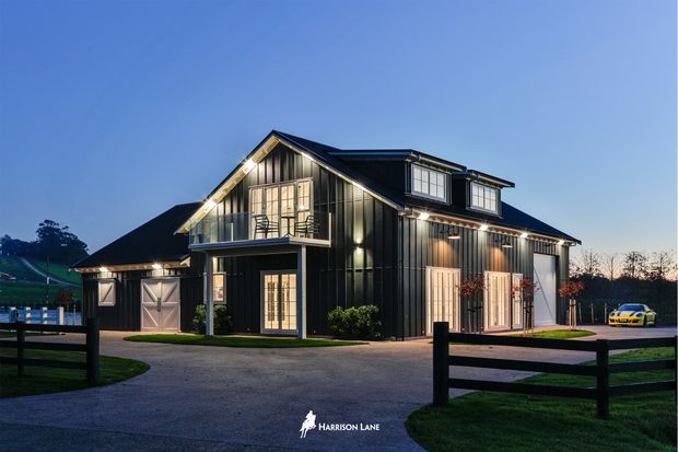 Exterior twilight photograph of a large dark-clad barn-style residence or rural property, featuring a prominent gabled main volume, dormer windows, covered balcony, vertical board-and-batten-style cladding, multiple garage or service doors, and a broad paved driveway. Fenced lawn and an adjacent smaller outbuilding are visible.