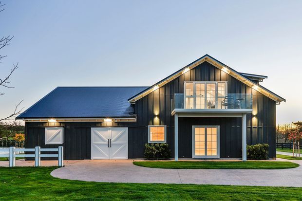 Exterior view of a dark-clad two-storey barn-inspired residence with a prominent gabled roof, attached garage, upper balcony, large glazed doors and windows, and a curved concrete driveway across a manicured lawn. The facade is photographed at dusk with warm interior and exterior lighting.