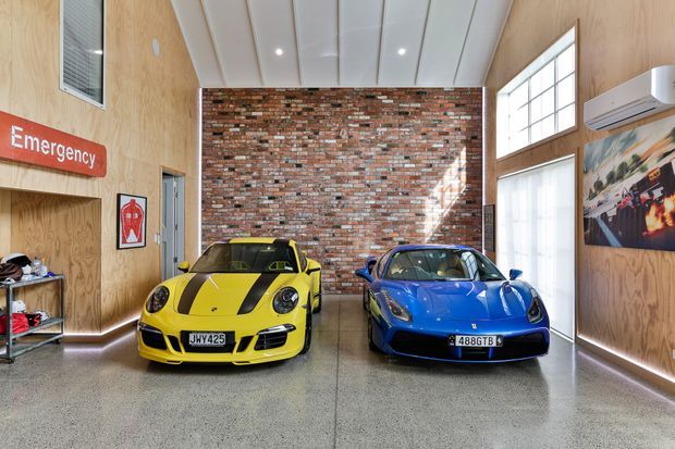 A bright, double-height automotive showroom or service space with two sports cars displayed in front of a red and multicoloured exposed brick feature wall. The room has warm plywood wall lining, polished concrete or terrazzo flooring, a white panelled ceiling with recessed lighting, high industrial-style windows, emergency signage, and visible automotive artwork.