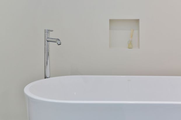 Close-up of a minimalist bathroom featuring a freestanding white bathtub with a tall chrome floor-mounted faucet, set against a plain light wall with a recessed niche.