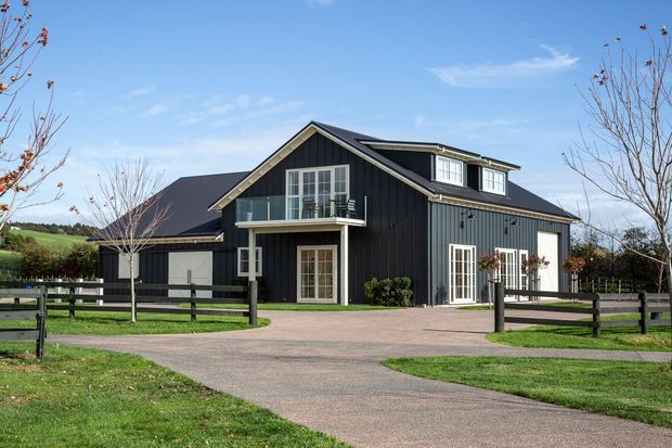 A dark-clad, multi-level rural house with a prominent front-facing gable, pitched roof, dormer window, balcony, white-framed glazing, and attached side wing. The home sits within a landscaped rural property with a broad paved driveway and timber post-and-rail fencing.
