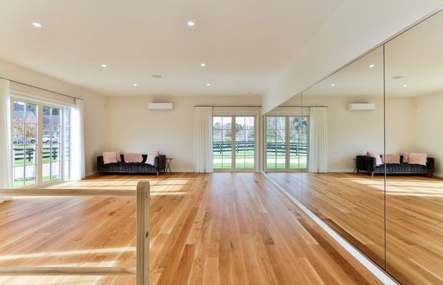 Bright, spacious mirrored studio room with polished timber flooring, cream walls, recessed ceiling lights, large glazed doors and windows, and a small dark sofa. The full-height mirrored wall reflects the room and outdoor greenery, creating an open and airy feel.