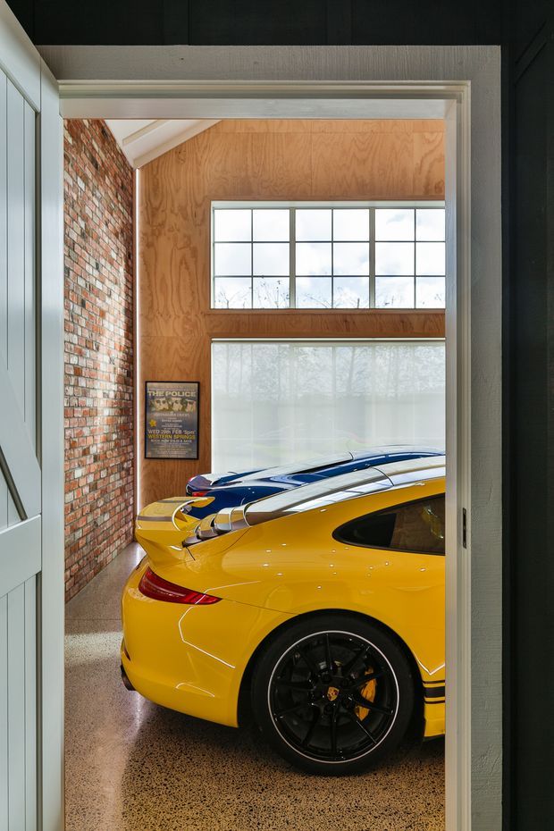 A view through a wide interior opening into a compact, high-end garage or car display space. A bright yellow sports car dominates the foreground, with a blue car behind it. The space combines exposed red brick, warm plywood wall lining, large industrial-style gridded windows, polished speckled concrete flooring, and dark-painted structural elements.