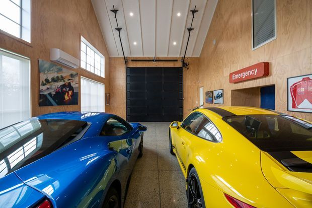 A bright, high-ceilinged automotive showroom or private car garage with polished concrete flooring, warm plywood wall lining, large clerestory windows, exposed ceiling services, framed automotive artwork, and two brightly coloured sports cars in the foreground. A large dark roller door and emergency signage are visible at the far end.