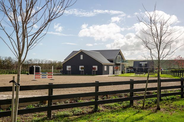 A rural property viewed from behind a dark timber post-and-rail fence. The main building is a dark-clad, gable-roofed house or barn-style structure with white trim, dormer-like upper glazing, and decorative planter boxes. The surrounding grounds include a sandy equestrian arena with colourful jump rails, young trees, hedging, and additional outbuildings under a bright sky.