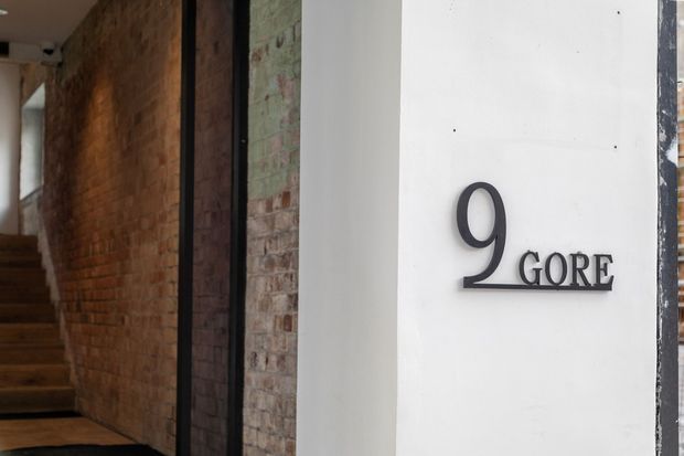 Close-up view of a building entry featuring a minimalist black address plaque reading “9 GORE” on a smooth white wall. An exposed red-brick wall, dark metal-framed glazing or doorway, and timber stairs leading inside are visible to the left, creating a contemporary industrial entrance aesthetic.