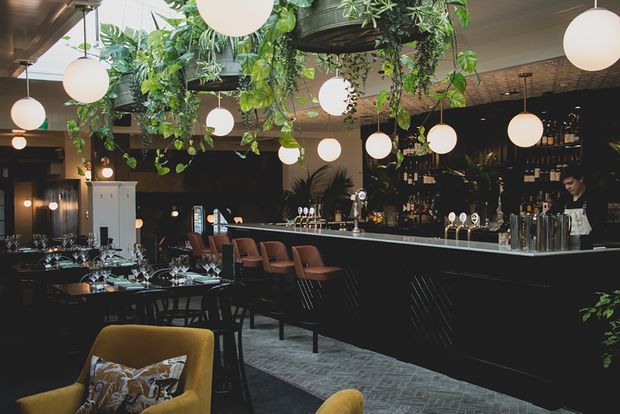 A moody, stylish commercial bar and dining venue with a long dark bar counter, back-bar liquor shelving, pendant globe lights, dining tables, upholstered bar stools, and abundant hanging greenery. The space has dark finishes, patterned flooring, and warm ambient lighting, creating an intimate hospitality atmosphere.