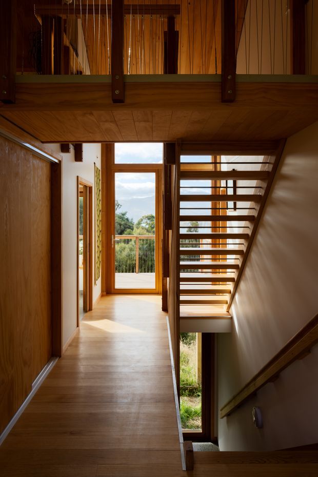 A warm timber-lined interior stairway and hallway. The view looks along a wood-floored passage toward a glazed door opening to a balcony or exterior deck, while an open-riser timber staircase rises on the right. A timber balustrade and upper-level void are visible overhead, with white plaster walls, extensive timber framing, and abundant natural light.