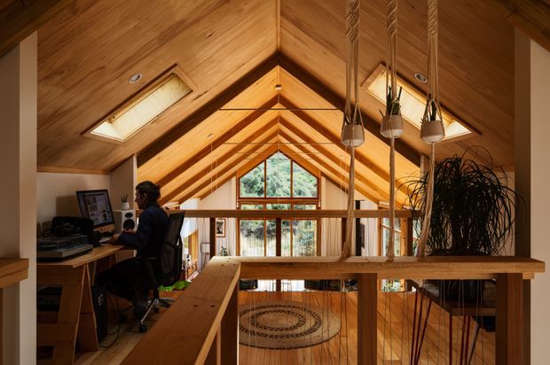 A warm timber-lined loft landing or mezzanine overlooking a double-height living space. The steep gabled ceiling has exposed timber rafters and two skylights, with a large triangular window at the far end. A compact built-in desk and computer workspace occupy the left side, while timber balustrades, hanging planters, and indoor greenery frame the open void.