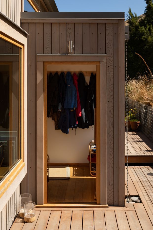 A compact timber-clad outdoor entry enclosure opens onto a small mudroom or coat-storage space. Vertical natural timber boards frame the doorway, with multiple coats hanging inside, timber flooring, and a raised timber deck continuing around the structure. A narrow walkway, chain, planter, and wire fencing are visible to the side, while strong sunlight creates warm shadows and highlights the natural wood grain.