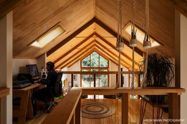 Warm timber-lined loft interior with a steep gabled ceiling, exposed structural rafters, skylights, and a large triangular window at the far end. A compact home office occupies the left side, while a timber mezzanine balustrade overlooks the lower level. Hanging planters and a circular rug add natural decorative elements.