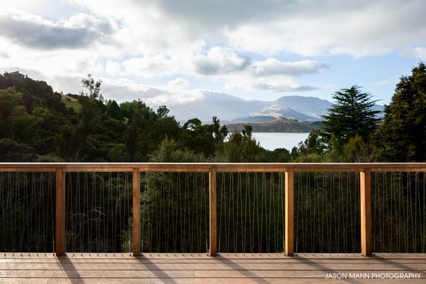 A timber deck or balcony overlooks dense native vegetation, a calm lake, and layered mountains beneath a partly cloudy sky. The foreground features a simple timber balustrade with slender vertical cable infill and broad timber decking, creating a scenic elevated outdoor viewing area.