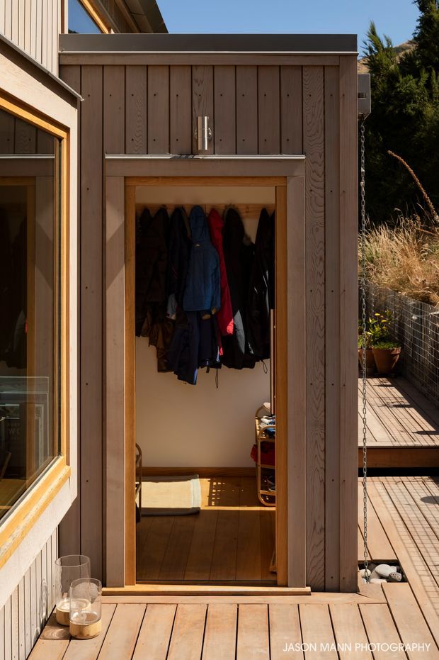 A sunlit compact timber-clad outdoor entry structure opens onto a small interior mudroom or storage space with coats hanging on hooks, timber flooring, and a narrow shelving unit. The surrounding deck and adjoining timber facade feature warm vertical wood boards, while a chain and wire railing frame the right side. The image has a warm, natural architectural photography style.