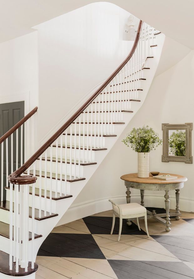Bright, refined residential stair hall featuring a sweeping curved staircase with white-painted balusters, dark timber handrails and stair treads, and pale walls. A round distressed console table with turned legs, upholstered stool, framed mirror, and floral arrangement sits beneath the stairs. The floor has a striking geometric pattern in dark and pale timber tones.