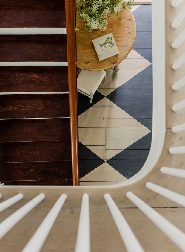 Top-down view through a curved stair or landing opening, showing dark stained timber stairs with white riser accents, pale timber flooring arranged in a bold black-and-natural geometric pattern, and a round timber table with a vase of greenery and a book below. White balustrade spindles frame the foreground and right edge.