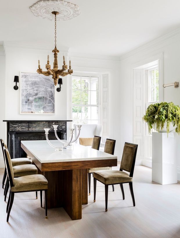 Bright, elegant dining room with a large white stone-topped dining table, dark wood pedestal base, upholstered dining chairs, and a black marble fireplace. The room features tall multi-pane windows with white paneled shutters, ornate plaster ceiling detailing, a traditional brass chandelier, white walls, and a hanging arrangement of greenery.