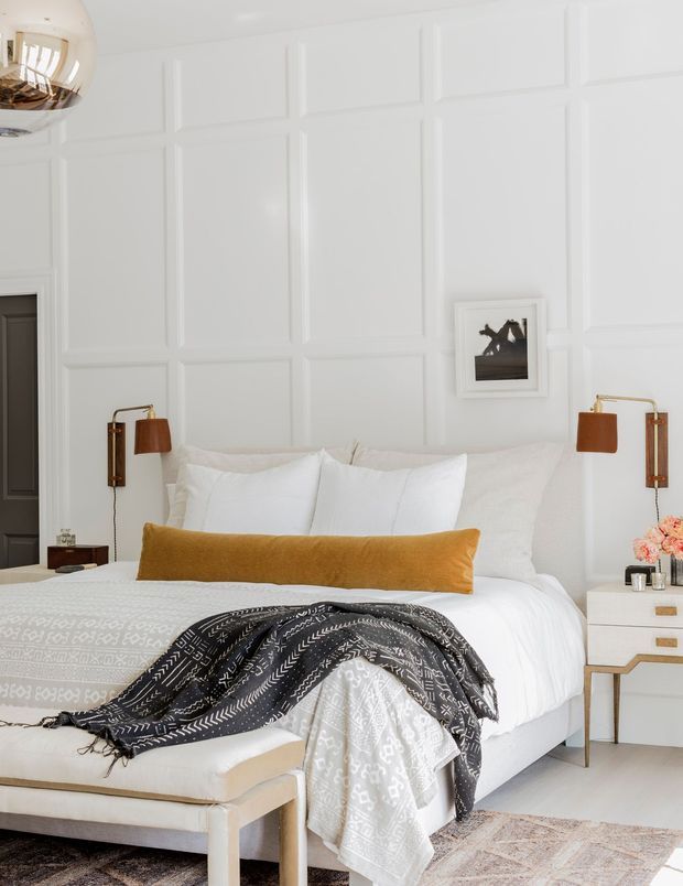 Bright, refined bedroom with a white upholstered bed dressed in layered white and patterned bedding, a mustard-gold lumbar cushion, and a dark patterned throw. The walls feature prominent white decorative panel moulding, complemented by brass-and-leather wall sconces, white bedside furniture with brass pulls, and a pale upholstered bench at the foot of the bed. A small framed artwork and partial mirrored reflection add detail.