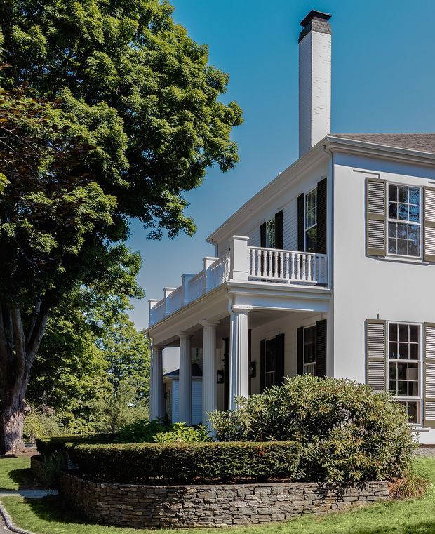 Exterior view of a stately two-storey white-painted house with a gabled roof, tall brick chimney, classical columns, upper-level balcony with turned balusters, decorative cornice, and louvered shutters. The home is set within mature trees and formal clipped hedges, with a low rough-stone retaining wall in the foreground.