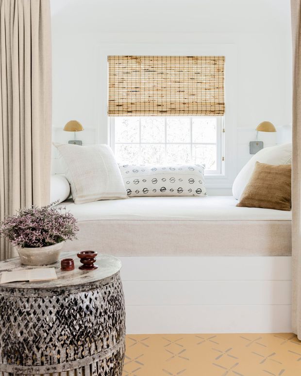 Bright, refined window-seat nook with a built-in white daybed beneath a large window. The seat is layered with white and beige cushions, patterned bolster pillows, and a pale upholstered front. Full-height light curtains frame the opening, while a woven bamboo Roman blind filters the daylight. A small round textured side table holds books, ceramics, and a potted plant, and the foreground has a warm patterned floor covering.