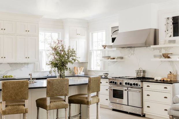 Bright, spacious white kitchen with traditional Shaker-style cabinetry, marble-look countertops and backsplash, a large island with three upholstered stools, open shelving, and a freestanding stainless-steel range beneath a sculptural rangehood. Abundant windows create an airy, refined atmosphere.