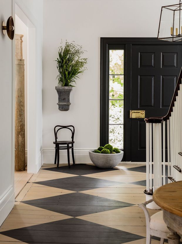 A refined residential entry hall with a black paneled front door, glazed sidelights, white walls and trim, a decorative wall planter, small chair, sculptural bowl, and a striking geometric painted timber floor in black and natural wood tones. A staircase with white balusters rises on the right, while a traditional lantern pendant and brass hardware add classic detail.