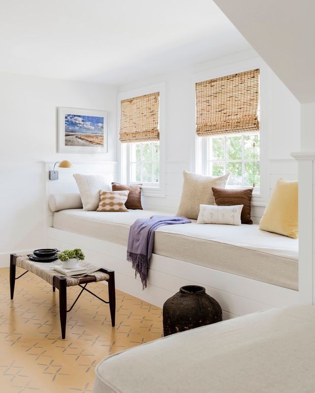 Bright, serene sitting room with two built-in window seats beneath tall divided-light windows. White painted wall panelling and trim are paired with woven bamboo blinds, neutral upholstered cushions, a patterned ochre tile floor, and a simple dark wood-and-metal coffee table. The room has abundant natural light and a relaxed coastal-inspired feel.