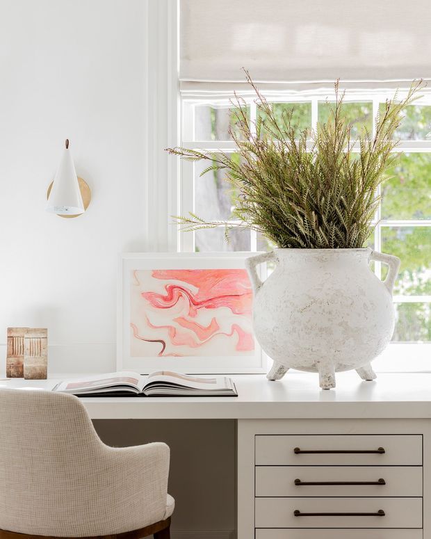 Bright, refined home office vignette featuring a white built-in desk with drawer storage, a textured upholstered chair, a large ceramic urn with branches, framed abstract artwork, and a wall-mounted sculptural light. A large window with a light roller shade fills the space with natural light.