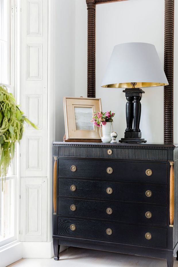 A refined interior vignette featuring a tall black chest of drawers with brass pulls and side accents, positioned beneath a large ornate dark wood-framed mirror. A black table lamp with a white shade and brass interior sits on top alongside a framed artwork and small floral arrangement. The setting includes bright white decorative wall panelling and a tall window, creating a classic, elegant atmosphere.