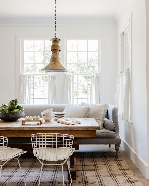 Bright, refined dining and living area with a rustic timber dining table, white wire-frame chairs, and a tailored grey sofa. Large multi-pane windows with sheer curtains fill the room with natural light, while a prominent aged brass pendant hangs above the table. A neutral plaid rug, white walls and trim, and soft linen cushions create a relaxed classic interior.