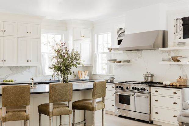 Bright, spacious traditional-style kitchen with white raised-panel cabinetry, marble-look surfaces, open display shelving, a large stainless steel range cooker and hood, and a central island with three upholstered stools. Large windows provide abundant natural light, while greenery and neutral furnishings add warmth.