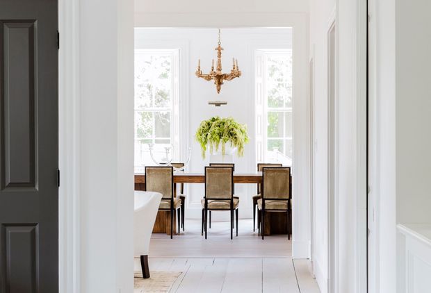 View through a bright white-trimmed hallway into a formal dining room. The dining area features a rectangular dark wood table, upholstered dining chairs, a central leafy plant, tall windows, and an ornate brass chandelier. Dark paneled door and white wall panelling frame the foreground.