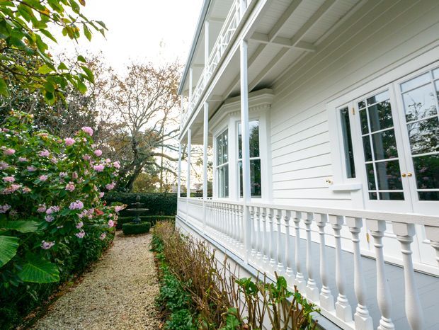 Exterior view of a white weatherboard house with a long covered verandah, decorative balustrade, white-framed French doors and windows, and a gravel garden path bordered by flowering shrubs and formal planting.