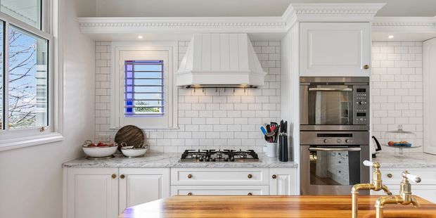 Bright, refined white kitchen with shaker-style cabinetry, ornate crown moulding, a large custom rangehood, white subway-tile splashback, marble-look benchtops, integrated stainless-steel ovens, a gas cooktop, and a timber island with brass/gold tapware. A window provides natural light and views to trees outside.