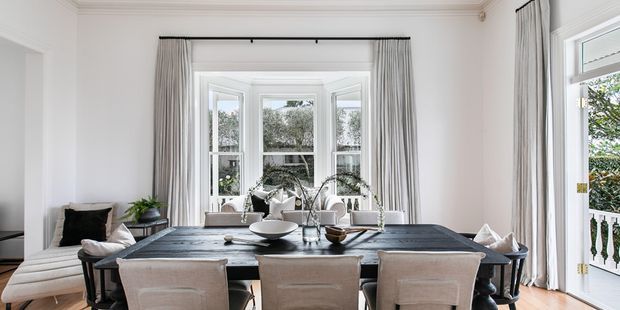 Bright, elegant dining room with a long dark dining table surrounded by upholstered chairs, set beneath a large bay window with full-height curtains. White walls, detailed cornice moulding, timber flooring, and French doors opening to a balcony create a refined traditional interior.