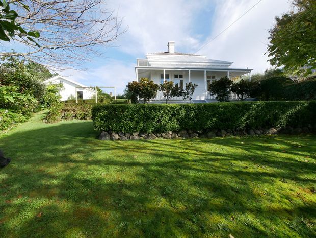 A traditional single-storey white weatherboard house viewed from a manicured garden. The home features a hipped roof, covered front veranda with slender posts and balustrading, white-framed windows, a central chimney, clipped hedges, and a simple lawn. A small white outbuilding or pergola is visible to the left.
