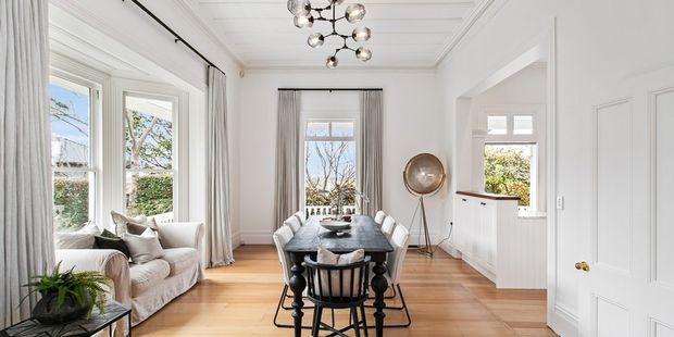 Bright, refined open-plan dining and living space with white decorative wall panelling, high detailed ceiling mouldings, tall windows dressed in sheer curtains, and warm timber flooring. A dark wood dining table with white upholstered chairs sits centrally, while a relaxed white sofa and greenery occupy the adjoining sitting area. Contemporary sculptural lighting and restrained furnishings create an elegant classic interior.