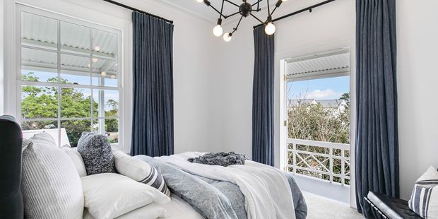 Bright, refined bedroom with white walls, a large bed dressed in layered white and grey bedding, tall dark blue curtains, expansive multi-pane windows, and a modern black branching pendant light. The room opens visually to leafy outdoor views and a balcony or terrace railing.