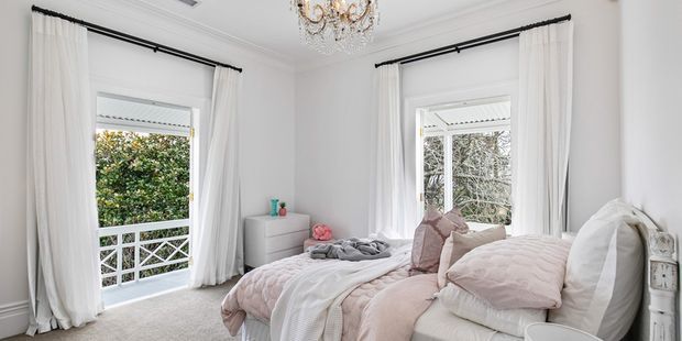 Bright, elegant bedroom with a large upholstered bed dressed in white and blush-pink bedding, white curtains on black rods, French doors opening to a balcony, a small white dresser, ornate chandelier, and detailed white wall moulding. The room has a soft, refined, airy character with a view of greenery outside.