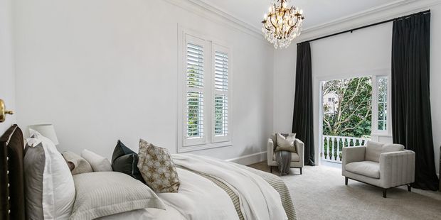 Elegant, light-filled bedroom with a large upholstered bed, layered white and neutral bedding, decorative cushions, a pair of armchairs, tall black curtains, plantation shutters, ornate cornice detailing, and a crystal chandelier. French doors open to a small balcony or terrace with greenery beyond.