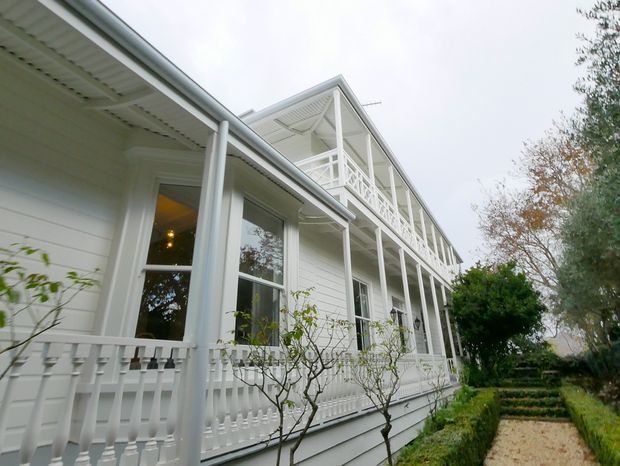 Exterior view of a large white timber-clad heritage-style house with an elevated wraparound verandah, decorative balustrades, slender posts, tall windows, and a second-storey balcony. A formal garden path and hedges run alongside the building.