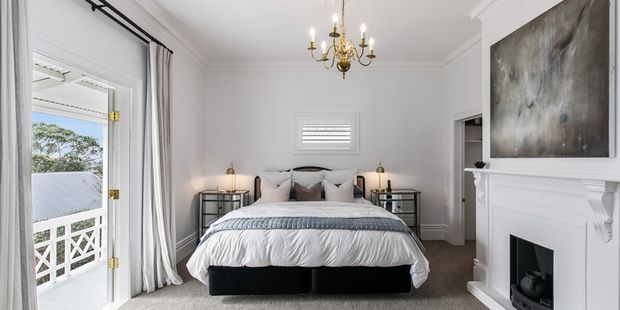 Bright, refined bedroom with a centered upholstered bed, layered neutral bedding, matching mirrored bedside tables, a classic white fireplace with mantel, ornate chandelier, plantation shutter, and full-height curtains opening to a small balcony. The room combines traditional architectural detailing with a calm contemporary-neutral scheme.