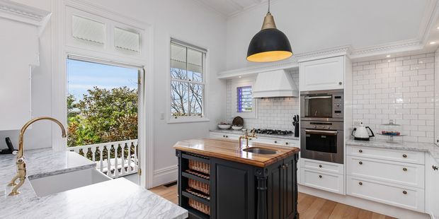Bright traditional kitchen with white paneled cabinetry, marble-look benchtops, a dark central island with timber countertop, subway-tile splashback, brass tapware, pendant lighting, and decorative moulding. A balcony or veranda is visible through the glazed door.