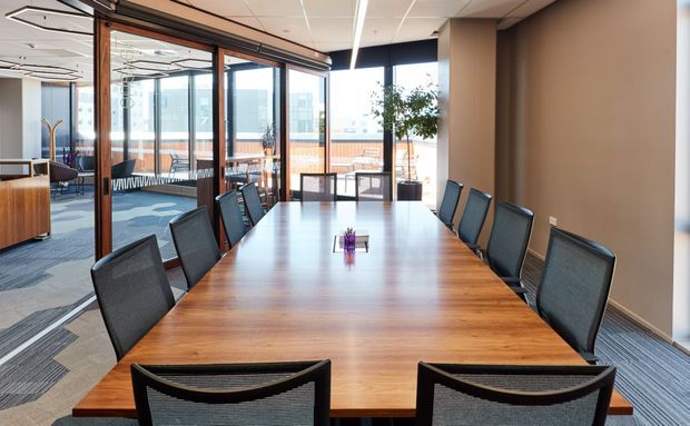A bright contemporary commercial meeting room with a long polished timber conference table surrounded by black mesh chairs. Full-height glazed partitions with dark timber frames separate the room from an adjacent open office, while expansive windows provide strong daylight and views of nearby buildings. The space has a neutral palette, carpeted flooring, suspended acoustic ceiling, and understated built-in finishes.