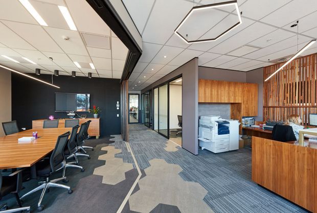 A contemporary commercial office interior featuring an open meeting room with a large timber conference table, black office chairs, glass-partitioned private offices, a reception or work counter, timber wall cladding and vertical timber screening. The space has patterned carpet flooring, a suspended tiled ceiling with linear and geometric pendant lighting, and a bright, professional fit-out.