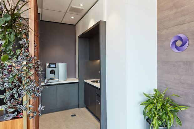 Compact contemporary kitchenette or office refreshment area with dark built-in cabinetry, a small benchtop coffee machine, integrated sink or work surface, and recessed shelving. The space has a light neutral floor, white walls, a grey feature wall, timber trim, recessed ceiling lights, and abundant indoor plants. A purple circular wall emblem is mounted on the adjacent wood-look wall.