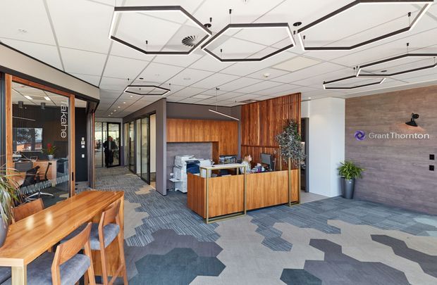 A contemporary commercial office reception and waiting area, identified by the Grant Thornton branding. The space features a timber reception desk and extensive vertical timber wall panelling, glazed meeting-room partitions, grey patterned carpet tiles, and a white suspended ceiling with geometric black-edged acoustic ceiling forms and recessed spot lighting. A small timber table and chairs occupy the foreground, with indoor plants and office equipment adding context.
