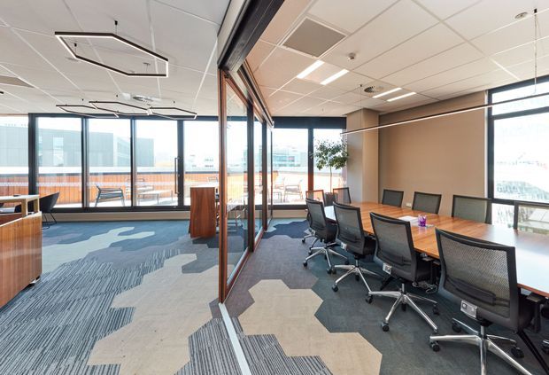 Bright contemporary commercial office interior with a large open-plan meeting and collaboration area. A long wood meeting table with black mesh office chairs occupies the right side, while glazed partitions separate it from an open workspace and balcony or terrace beyond. The space has patterned modular carpet tiles, warm wood joinery, full-height glazing, suspended linear lighting, and a suspended acoustic tile ceiling.