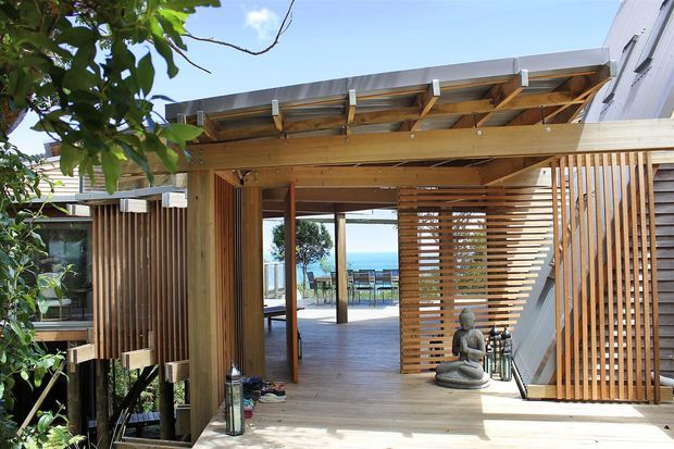 A timber-framed outdoor deck and covered terrace adjoining a modern building. The space features warm timber decking, exposed timber beams and rafters, vertical timber screening, horizontal slatted privacy panels, and open views toward a blue waterfront or sky beyond. Outdoor furniture and a small decorative figure are present, with lush planting at the edges.